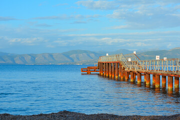 Pacific landscape of a rusted pier with bluish atmosphere, pacific sea and green mountains in the background in cloudy day. Kavos beach in Corfu Island, Greece.