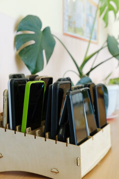 Mobile phones stored in a wooden box, organized neatly, with a green plant in the background, illustrating the concept of phone storage during school lessons