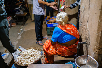 Elderly Indian woman in a vibrant orange saree selling fresh garlic at a traditional street market, capturing daily commerce and cultural urban life.