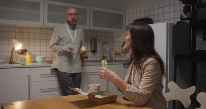 Side view girl in business clothes office worker holding sandwich in hands and talking with her colleague guy who leaned on kitchen counter During lunch in office kitchen