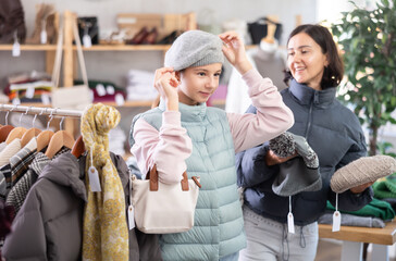 Obraz premium Teen girl is fitting on and trying on winter hat in store, family shopping with mom. Buyer looks at herself in mirror while shopping, evaluates her appearance. Fitting clothes and accessories.