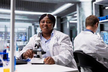 Portrait of black woman posing in laboratory at desk with microscope, examining specimen for a research study. Scientist in white coat emphasizing cellular discovery in bioscience.