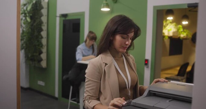 Office woman using printer or copier at workplace while colleague works in background business routine document printing in modern green coworking space