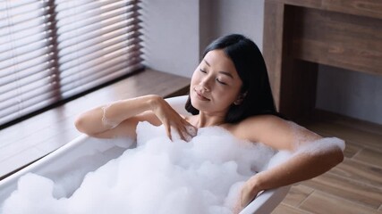 A young Asian woman enjoys a relaxing bubble bath at home, indulging in her daily skin care routine in a stylish bathroom filled with natural light, creating a calming ambiance. - Powered by Adobe