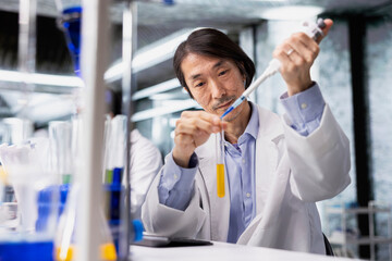 Senior scientist uses pipette to transfer liquid into test tube containing yellow solution at laboratory bench. Asian man does pipetting procedure with chemical liquid solution in test tube