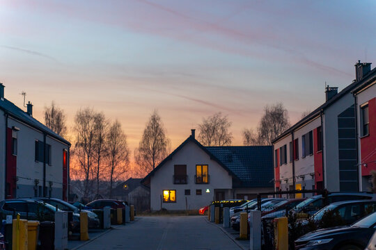 Peaceful winter evening in suburban residential neighborhood with colorful houses, warm glowing windows and beautiful pink purple sunset sky over bare trees. Cozy community lifestyle concept.