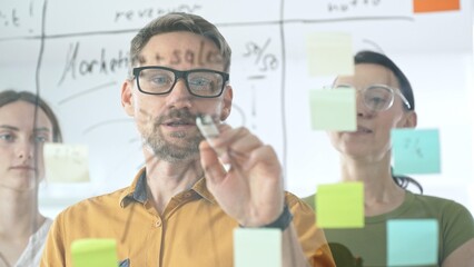 Professional team brainstorming project strategy, placing colorful sticky notes on glass board, analyzing data, sharing insights, developing collaborative solutions