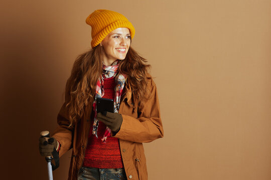 A smiling woman in warm outdoor attire, including a yellow beanie and red sweater, holds both a smartphone and a trekking pole, embodying readiness for adventure. - Powered by Adobe