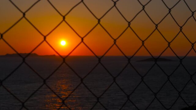 Border at sunset. Warm sunlight illuminates the sky and reflects on the calm ocean surface, viewed through a metal wire mesh