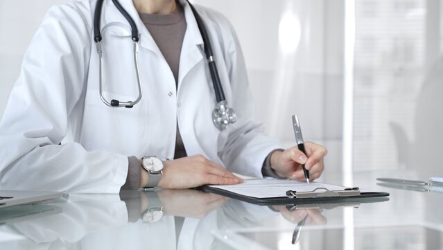 Female doctor in white coat with stethoscope writing patient notes on clipboard, completing medical records in clinic office. Medicine and health care