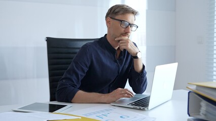 Naklejka premium Professional businessman sitting at a sleek desk, working on a laptop and thinking about goals in modern office filled with natural light. Business people concept