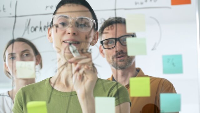 Professional team brainstorming project strategy, placing colorful sticky notes on glass board, analyzing data, sharing insights, developing collaborative solutions