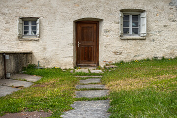 Front facade of a country house with a garden, wooden door, two windows of different sizes, and a stone path.