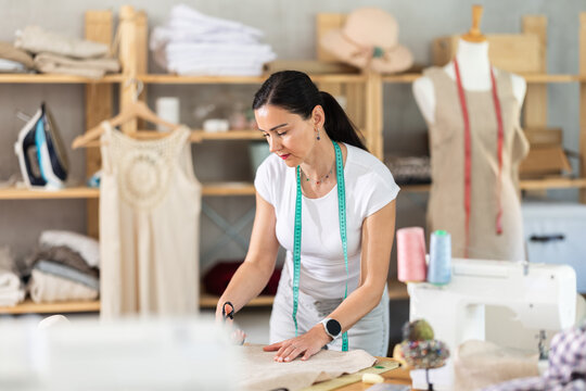 In workshop, woman dressmaker cuts fabric, cuts out fabric according to drawing. Craftsman cuts off pieces of fabric to create product, preparatory stage of work.. - Powered by Adobe