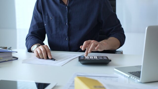 Professional businessman calculating financial data, reviewing documents near laptop in modern workplace setting. Audit and taxes, business concept - Powered by Adobe