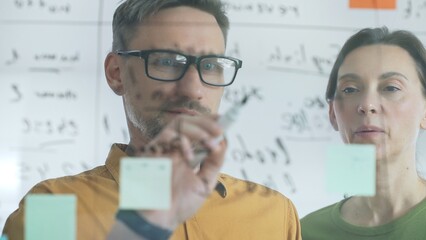 Professional team members brainstorming strategy, writing notes on transparent whiteboard with colorful sticky papers during collaborative meeting. Business people concept