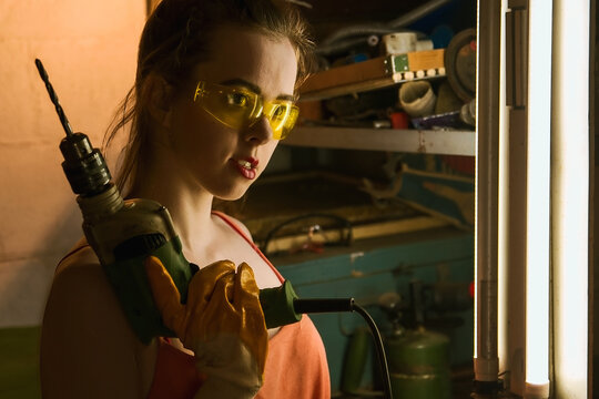 A female construction worker is holding a drill in her hand, wearing yellow safety glasses and yellow protective gloves, a tshirt, and makeup on her face, looking away.