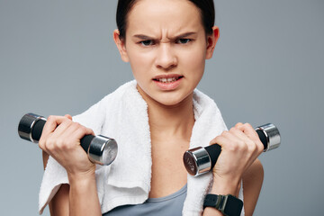 A focused young woman lifts chrome dumbbells with a white towel over her shoulders and a fitness...