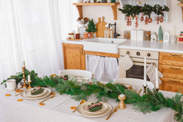 Elegant Christmas holiday table setting with greenery and simple decor on cozy kitchen background. Beautifully arranged holiday table with plates, candles and golden cultery on white tablecloth