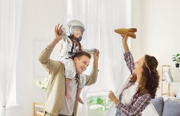 Child astronaut daughter having fun with mother and father. At home parents plays girl in costume and helmet rides dads shoulders while mom lifts rocket. Happy family space dream and imagination.