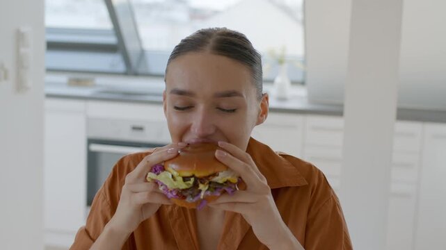 A young woman sits at a bright kitchen table, savoring a large burger filled with fresh ingredients.