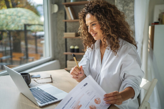 Fototapeta Woman working remotely with laptop analyzing home office paperwork