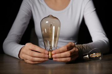 Young woman sitting by table with light bulb on dark background, closeup