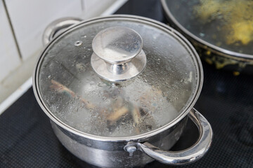 A stainless steel pot with a condensed glass lid sits on a black stovetop, cooking an unseen meal.