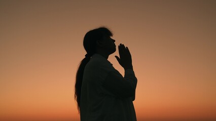 Christian woman praying at sunset, close-up. Girl against sky prays to God for family and children. Faith in God. Relaxation and meditation in nature. Faith Hope Love. Religion. Silhouette woman, sky