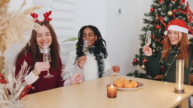 Three multiracial friends toast with glasses, surrounded by a festive atmosphere of a Christmas tree, garlands, gifts, and holiday treats on the table.