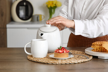 Young woman pouring hot tea from teapot into cup in kitchen, closeup