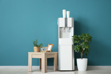 Modern water cooler, table and houseplant near blue wall in room