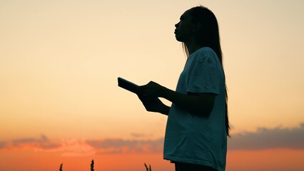 Young woman reads book in autumn park in rays of sunset. Prayer in nature. Student reads textbook in nature in spring. Man reading book outdoors. Studying, education. Beautiful woman praying at sunset