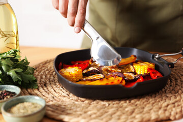 Man taking tasty grilled vegetables from pan with tongs at wooden table indoors, closeup