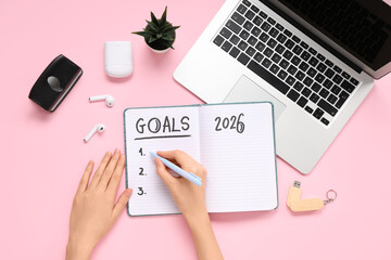 Female hands writing 2026 goals in notebook with laptop on pink background