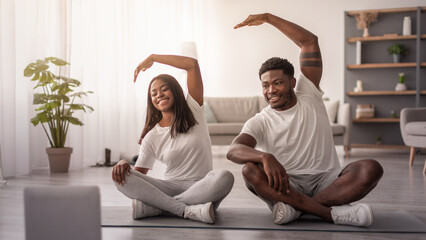 Happy African American Couple Doing Side Bend Stretch Exercise, Working Out Together In Living Room At Home, Using Pc, Sitting On Floor Yoga Mat Looking At Screen. Fitness Workout, Domestic Training
