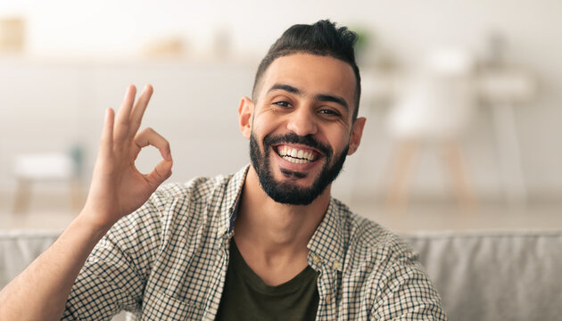 A man with a beard is smiling broadly while making an okay hand gesture. He is sitting in a relaxed position indoors, surrounded by a cozy living room atmosphere filled with soft light.