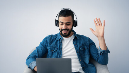 A young man in a denim shirt smiles and waves while participating in an online chat. He sits comfortably on a chair with a laptop in front of him, showcasing a relaxed atmosphere.