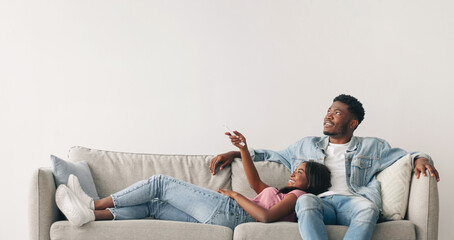 Cheerful Black Lady Holding Remote Controller, Opening Air Conditioner At Home While Relaxing On...