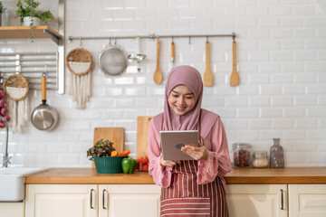 Smiling muslim woman wearing hijab use digital tablet in kitchen while preparing healthy food, online recipe search, home cooking and technology, halal cooking content, food blogger, healthy lifestyle