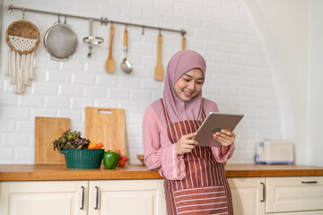 Smiling muslim woman wearing hijab use digital tablet in kitchen while preparing healthy food, online recipe search, home cooking and technology, halal cooking content, food blogger, healthy lifestyle