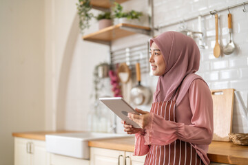 Smiling muslim woman wearing hijab use digital tablet in kitchen while preparing healthy food, online recipe search, home cooking and technology, halal cooking content, food blogger, healthy lifestyle