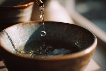 Close up of water dripping from metal faucet into ceramic bowl, sunlight creates soft reflections and tranquil atmosphere, highlighting texture and calm mood