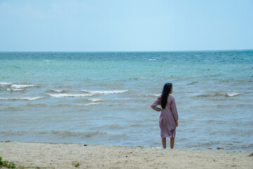 long haired asian woman wearing light purple clothes with long sleeves standing facing the sea on the beach with bright sky background for advertis