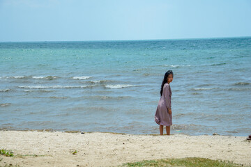 long haired asian woman wearing light purple clothes with long sleeves standing facing the sea on the beach with bright sky background for advertis