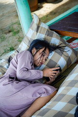 A view of an Asian woman with long black hair lying on a checkered patterned pillow while holding a cellphone with both hands and with a serious expre