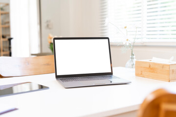 A modern laptop with a blank white screen on a clean white desk, perfect for digital mockups, against a bright, sunlit window background.