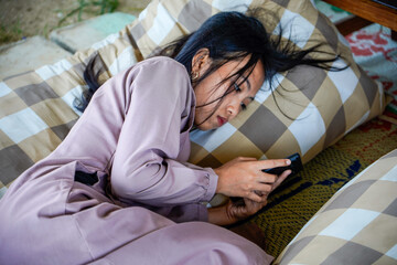 A view of an Asian woman with long black hair lying on a checkered patterned pillow while holding a cellphone with both hands and with a serious expre