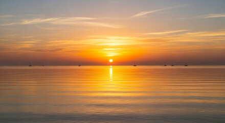 Obraz premium Golden hour paints the tranquil sea with vibrant hues as the sun dips below the horizon, casting a warm glow on the calm waters and distant fishing boats