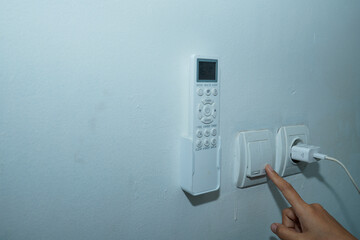 hand trying to turn on the light switch on the white wall with the AC remote control on the left and the socket with the charger cable connected to the right.